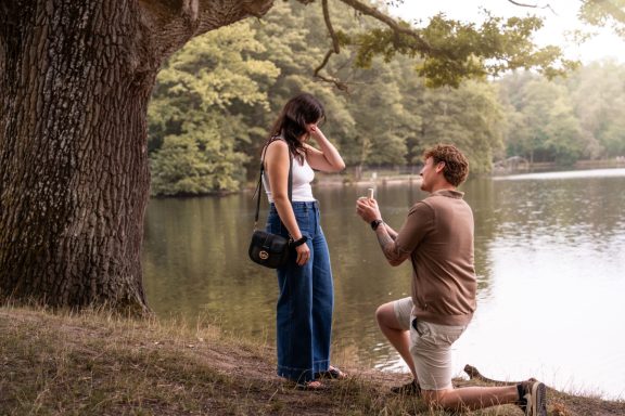 Romantischer Heiratsantrag am See mit kniendem Mann und ĂĽberraschter Partnerin in natĂĽrlicher Umgebung.